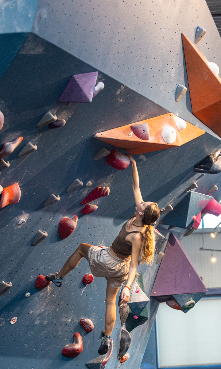 A woman climbs an indoor bouldering wall at an Austin climbing gym, reaching up with her right arm to grip a large orange hold amid colorful geometric grips. She wears a brown tank top and white shorts.