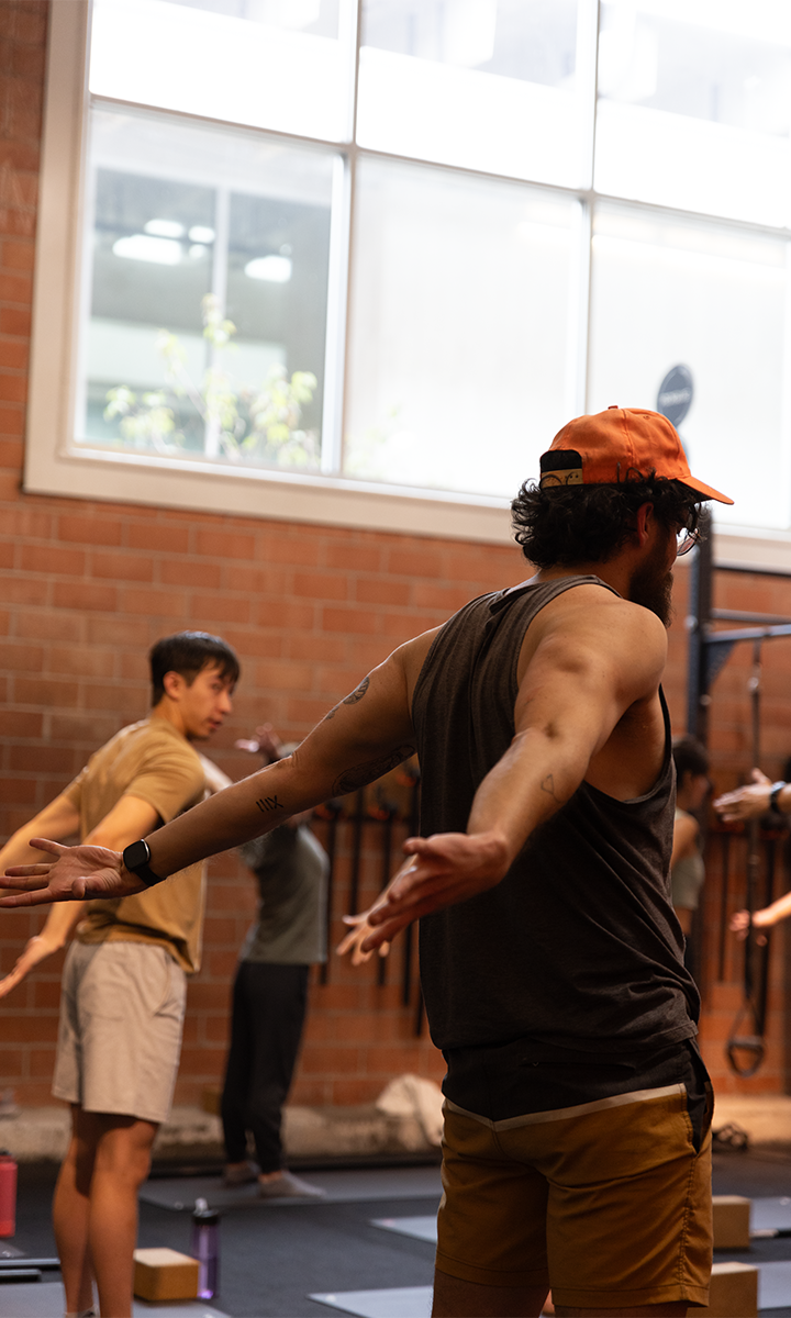 A group of people in athletic wear participate in a fitness or yoga class indoors at an Austin climbing gym. A man in the foreground, wearing a cap and tank top, stretches his arms out wide, with others following along in the background.