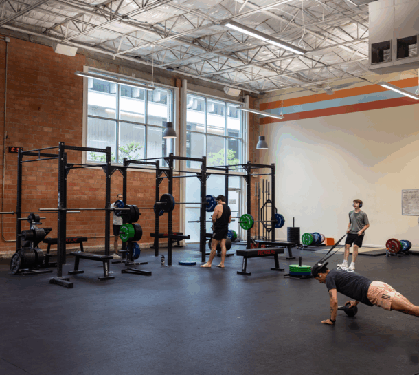 A spacious gym with large windows, several weightlifting racks, and gym equipment. Four people are present: one lifting weights, one observing, one adjusting equipment, and one doing push-ups on the floor at this Austin climbing gym.
