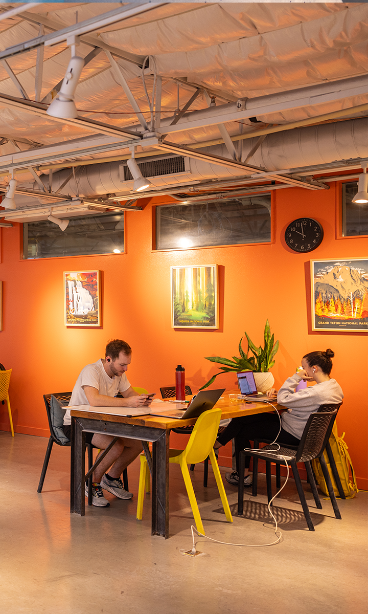 Two people work at a table in a brightly lit room with orange walls, colorful paintings, and a black clock—reminiscent of an Austin climbing gym. One uses a laptop while the other writes; indoor plants and exposed ceiling pipes complete the scene.