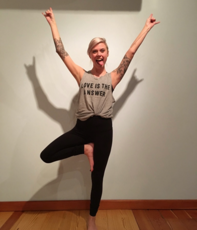 A person with tattoos stands in a yoga tree pose on a wooden floor, arms raised and fingers making rock signs, wearing a gray tank top that says LOVE IS THE ANSWER, smiling and sticking their tongue out.