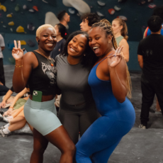 Three women pose smiling and flashing peace signs at Brooklyn Climbing Yoga and Fitness, surrounded by other people. They wear athletic outfits and appear happy and energetic, with climbing holds visible on the wall in the background.