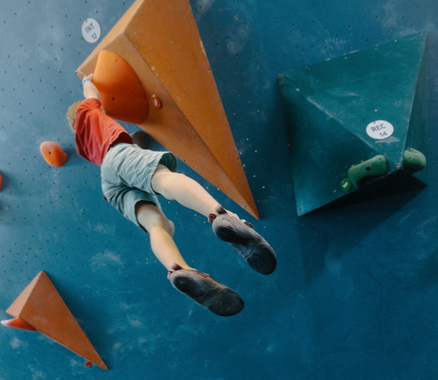 A person wearing a red shirt and green shorts climbs an indoor bouldering wall, reaching for an orange triangular hold with both hands—a scene reminiscent of Competitive Youth Climbing Teams in Austin training hard.