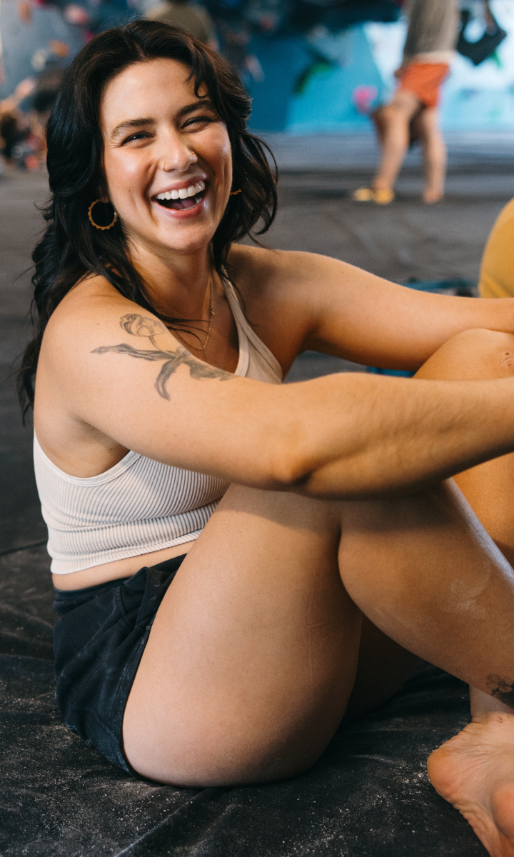 A smiling woman with dark hair, wearing a white tank top and black shorts, sits relaxed on the floor at Climbing Tempe. With a tattoo on her left arm, she appears happy as people and colorful climbing holds fill the background.