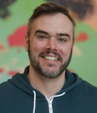A smiling man with short brown hair and a beard, wearing a dark green hoodie with white drawstrings, stands in front of a blurred colorful background in Minneapolis, ready to offer expert personal training.