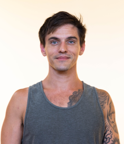 A man with short brown hair wearing a gray tank top stands against a light background, smiling slightly. He has a visible tattoo on his left shoulder and chest, looking ready for an afternoon at the Austin climbing gym.