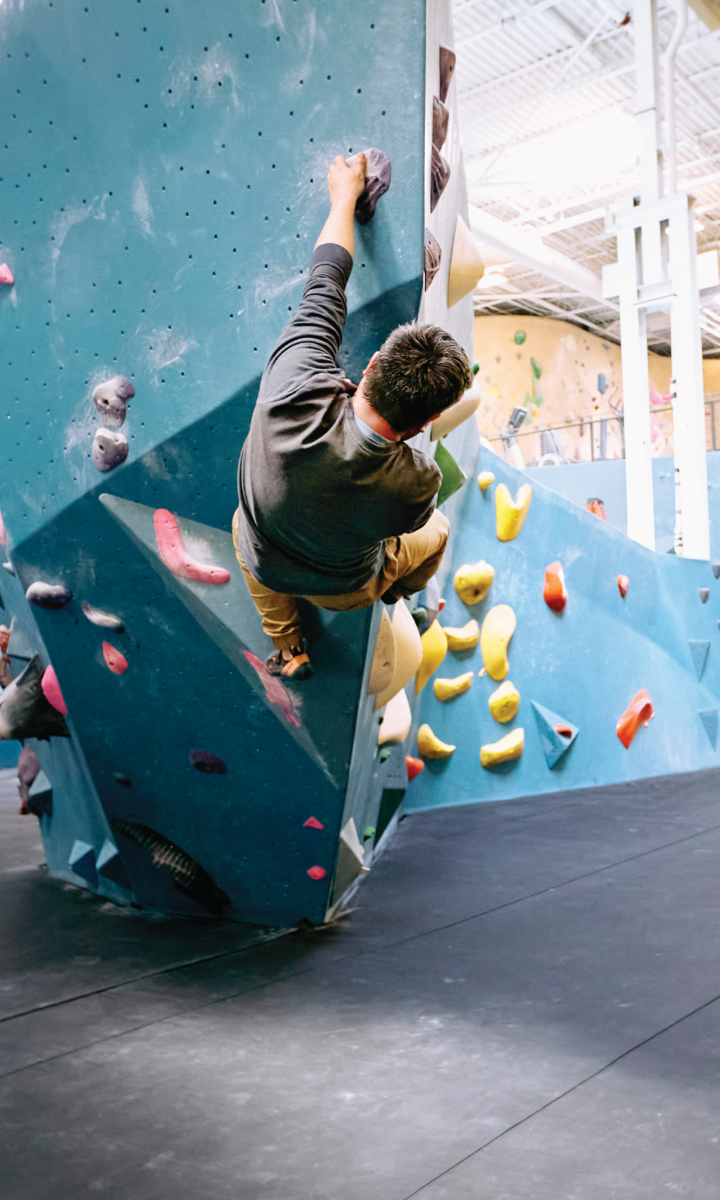 A person climbs an indoor rock wall, gripping colorful handholds with one arm extended, while wearing casual athletic clothing. The gym has a padded floor and bright lighting.