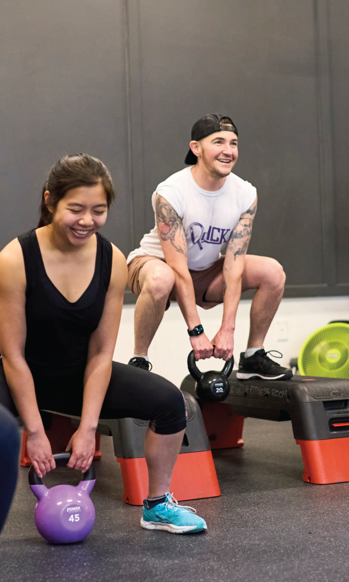 Two people exercising in a gym, both smiling and holding kettlebells while squatting on steps. The woman in front has a purple kettlebell; the man behind wears a cap and holds a black kettlebell.