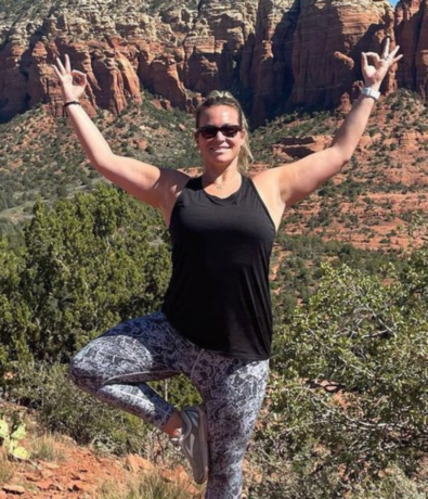 A woman in sunglasses and workout clothes balances in a yoga tree pose outdoors, smiling with arms raised, against a backdrop of red rock formations and greenery under clear sunlight.