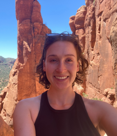 A woman smiling and taking a selfie outdoors in front of tall red rock formations under a bright blue sky. She is wearing a sleeveless black top and sunglasses on her head.