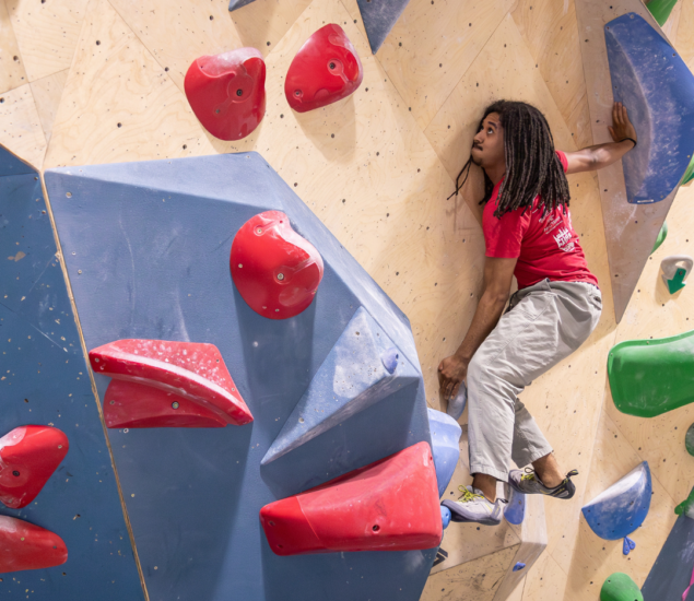 A person with long hair, wearing a red shirt and light pants, climbs an indoor bouldering wall at Eckington. They grip the wall with both hands and look up, appearing focused among red, blue, and green holds—perfect for the Partners Page.