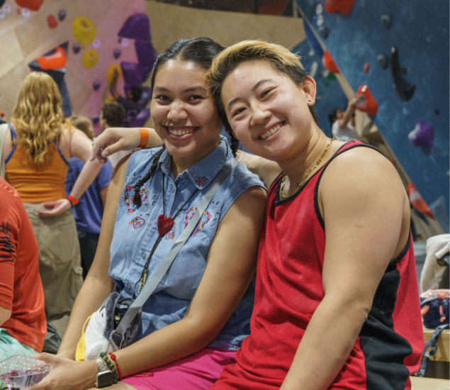 Two people sit closely together, smiling at the camera in a lively Eckington indoor rock climbing gym, with colorful climbing holds and other climbers in the background.