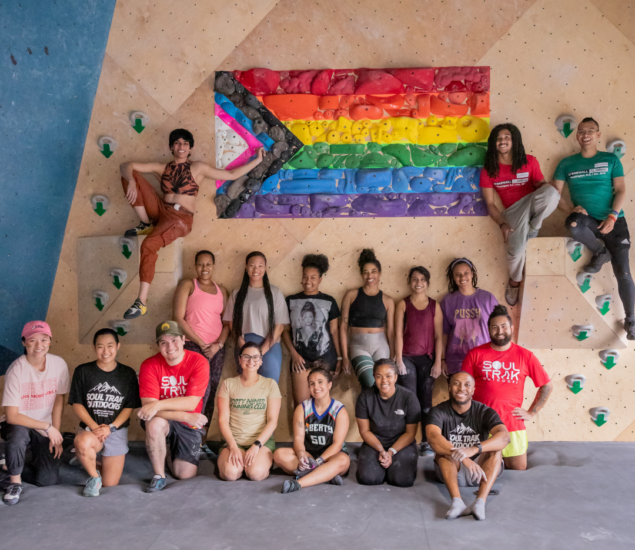 A diverse group of people pose and smile in front of a climbing wall at Eckington, featuring a large Progress Pride flag made of colored holds. Some are sitting on the ground, others are sitting or standing on the climbing wall.
