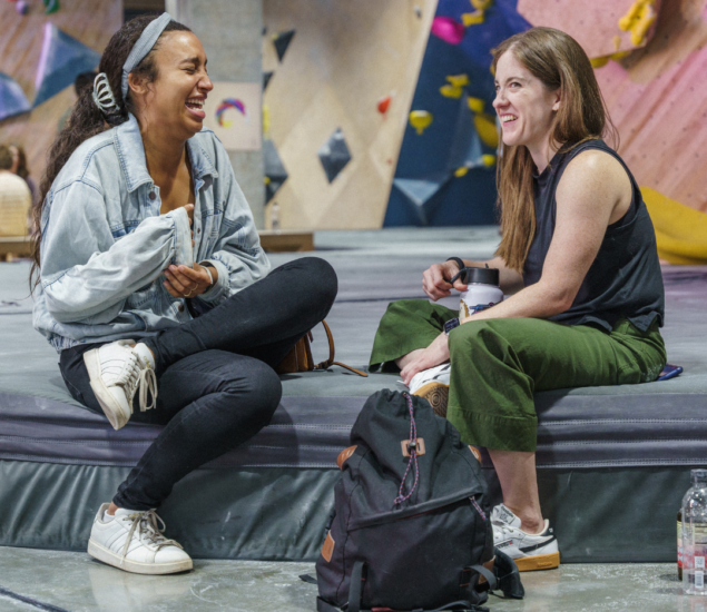 Two women sit on a mat at an indoor climbing gym in Eckington, smiling and talking. Dressed casually with backpacks and water bottles nearby, they enjoy the vibrant atmosphere as colorful climbing walls fill the background.