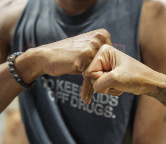 Two people give each other a fist bump—one wears a beaded bracelet, the other has a geometric tattoo. In the background, someone in a sleeveless shirt with partially visible text stands.