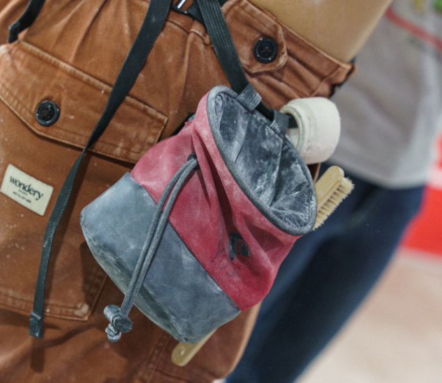 A close-up of a person wearing brown pants with a red and gray chalk bag, a brush, and tape attached to their belt, commonly seen on the Eckington Partners Page for rock climbing enthusiasts.