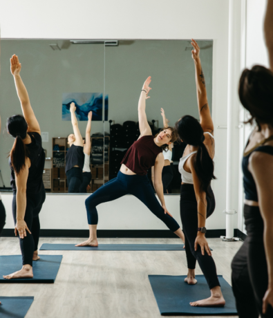 A group of people in athletic wear practice yoga poses on mats in a bright studio, following an instructor positioned in front of a large wall mirror.