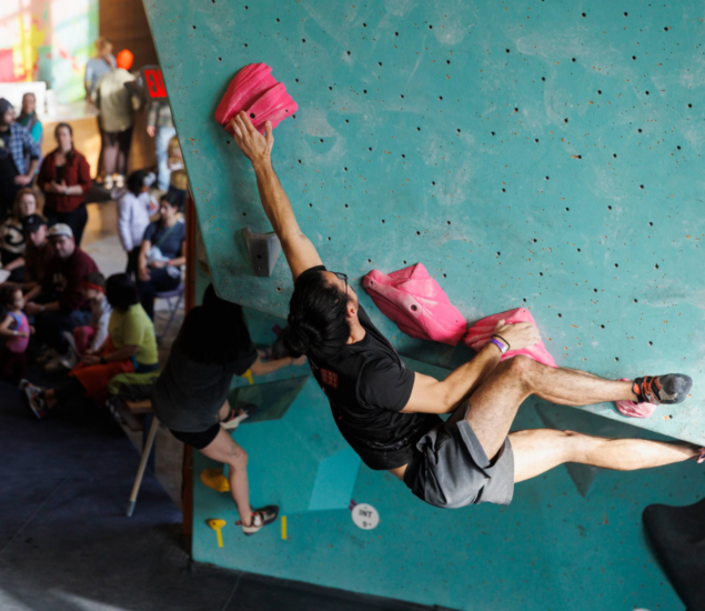 A man in the Adult Climbing League scales an indoor bouldering wall using bright pink holds, stretching his body horizontally. People in the background watch and sit, creating a lively atmosphere.