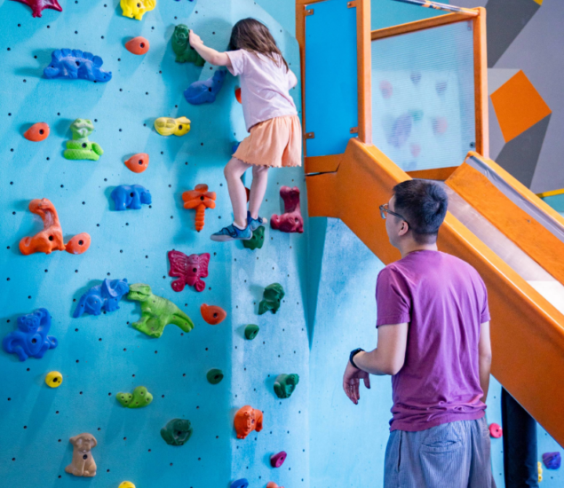 A young girl climbs a colorful indoor rock climbing wall while an adult man stands nearby, watching and supervising her. The wall is blue with various brightly colored holds, next to an orange slide.