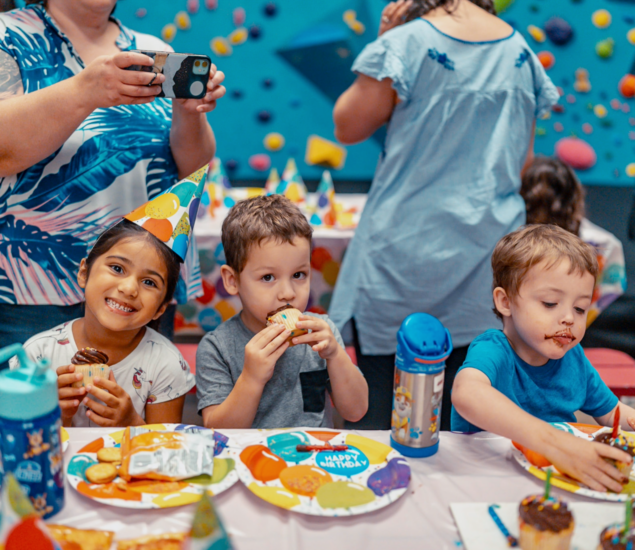 Three young children sit at a colorful table eating cupcakes at a birthday party, wearing party hats and smiling. Adults stand behind them, one taking photos. The scene showcases the fun found in Minneapolis Youth Programs. A blue climbing wall adds excitement to the background.