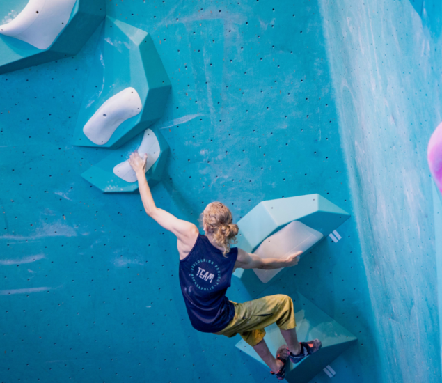 A person with blonde hair in a ponytail climbs a blue indoor bouldering wall, reaching for a white hold, dressed in a navy tank top and yellow pants—part of 2025 Youth Programs Minneapolis.