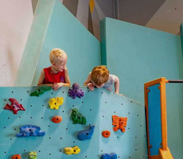 Two young children lean over a turquoise indoor climbing wall decorated with colorful animal-shaped holds, smiling and looking down. The playful scene could be part of Minneapolis Youth Programs in a vibrant indoor playground.