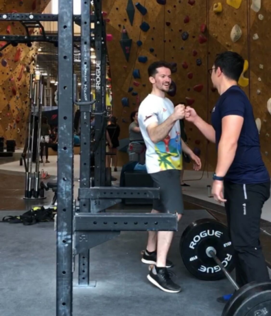 Two men are fist bumping and smiling next to a barbell in a gym with rock climbing walls in the background. Other people and gym equipment are visible, creating an energetic and social atmosphere.