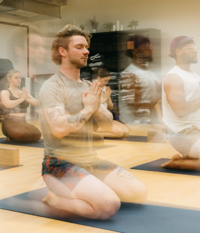 A group of people practice yoga indoors, kneeling on mats with hands pressed together in prayer position. The image has a motion blur effect, creating a sense of calm movement and focus.