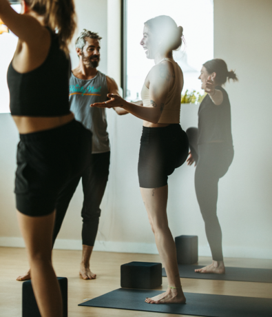 Three people in athletic wear are standing on yoga mats in a bright room, smiling and talking. One person gestures with their hands while another points, and yoga blocks are on the floor nearby.