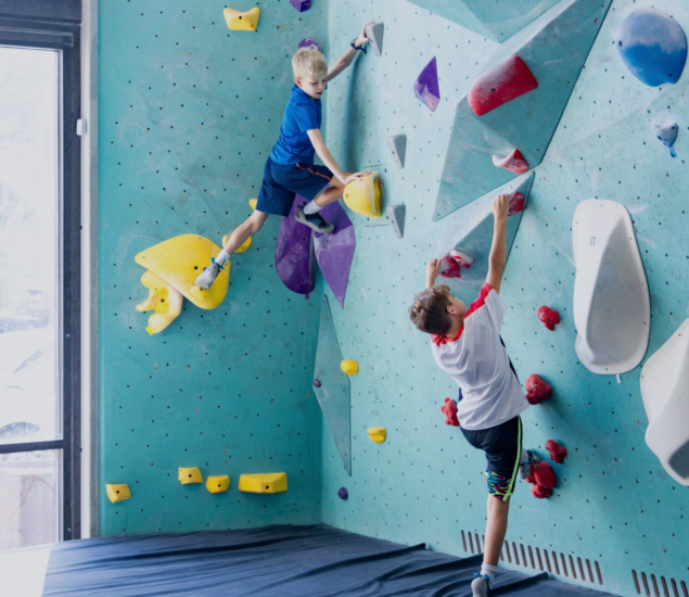 Two young boys are climbing a blue indoor bouldering wall with colorful holds during an After School Programs Minneapolis session. One boy in blue climbs higher, while the other in a white and red shirt is below him. A padded mat covers the floor.