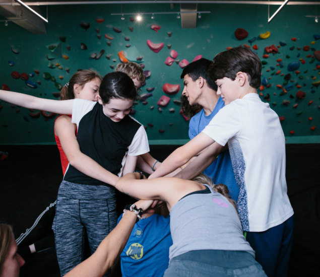 A group of young people stand in a circle, reaching into the center with their hands stacked together, smiling and interacting, with a colorful indoor climbing wall in the background—perfect for Youth Programs activities.