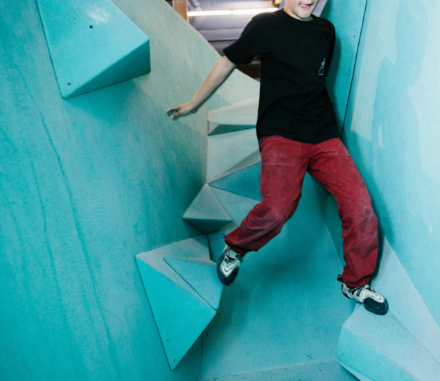 A person in a black shirt and red pants climbs between two blue walls using triangular climbing holds in an indoor gym featuring various youth programs.