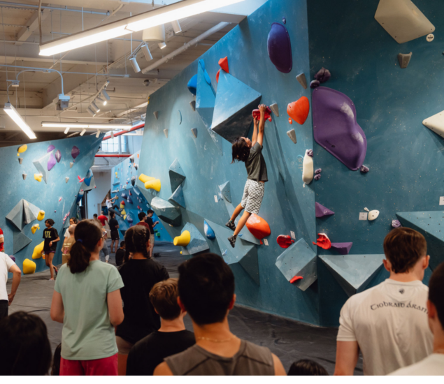 A group of people in a climbing gym watch as someone scales a blue indoor bouldering wall with colorful holds. The climber, gripping red holds, showcases the excitement often found during Youth Programs while others observe from the ground.