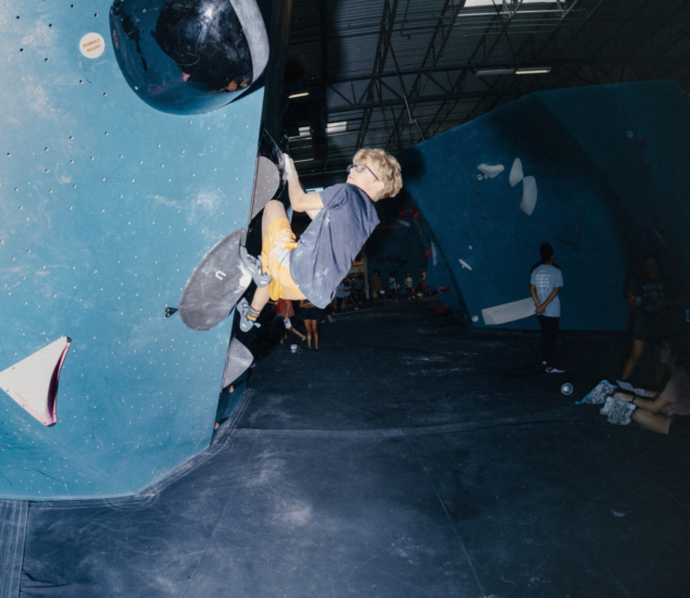 A person wearing glasses and orange shorts climbs a steep indoor bouldering wall, gripping black holds, as onlookers from Brooklyn youth programs watch alongside other climbing walls in the background.