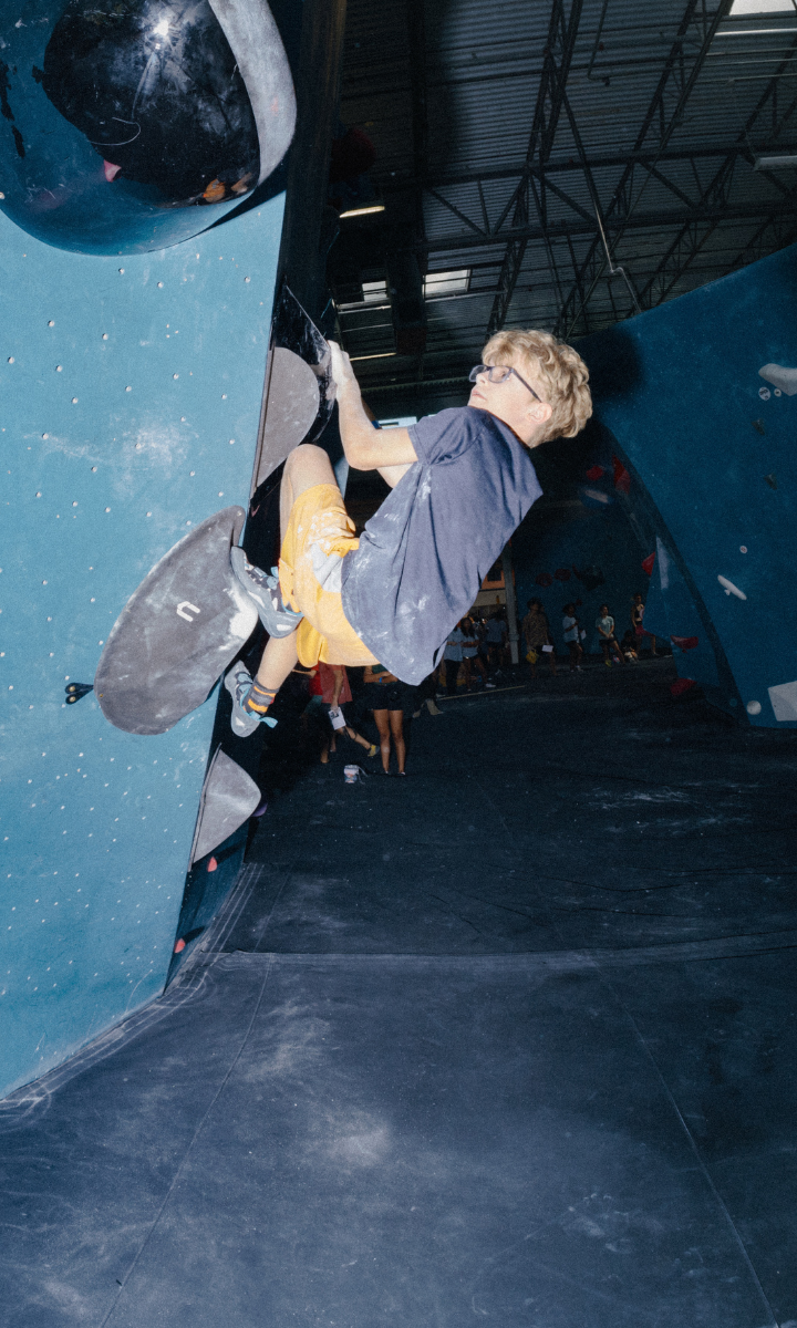 A young boy wearing glasses and orange shorts climbs a blue indoor bouldering wall at a Brooklyn gym, gripping large holds. Youth teams practice nearby, while the ceiling and other climbers are visible in the background.