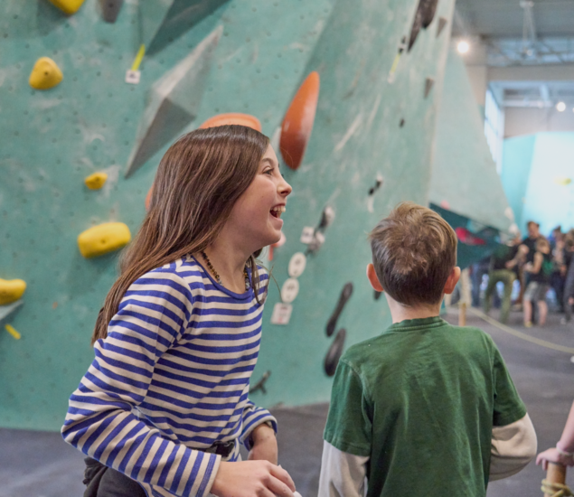 Two children stand in front of an indoor climbing wall, enjoying Minneapolis After School Programs. The girl in a blue and white striped shirt is smiling and laughing, while the boy in green faces away. Other people are in the background.